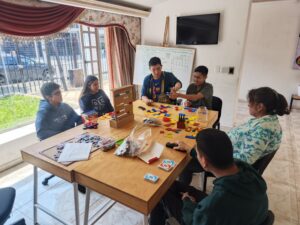 Grupo de jóvenes y una facilitadora sentados alrededor de mesas de madera compartiendo y participando en un taller dinámico de estimulación cognitiva con diversos juegos de mesa, cartas y materiales didácticos.