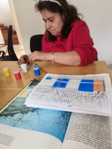 Una mujer concentrada pinta un dibujo en un cuaderno cuadriculado utilizando témperas de colores durante un taller de arteterapia y estimulación cognitiva.