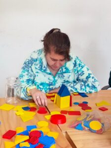 Una joven concentrada organiza y construye una pequeña estructura utilizando bloques lógicos de plástico de colores primarios (amarillo, azul y rojo) sobre una mesa de madera durante una actividad de desarrollo cognitivo.