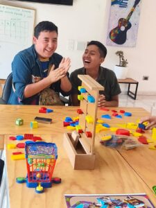Dos jóvenes sonríen y aplauden entusiasmados sentados frente a una mesa de madera con diversos juegos didácticos, bloques geométricos de colores y materiales de estimulación cognitiva.