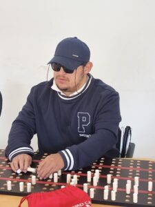 Joven con discapacidad visual utiliza un juego de mesa adaptado (tiflológico), manipulando con sus manos piezas cilíndricas de madera sobre un tablero táctil diseñado con relieves y agujeros.