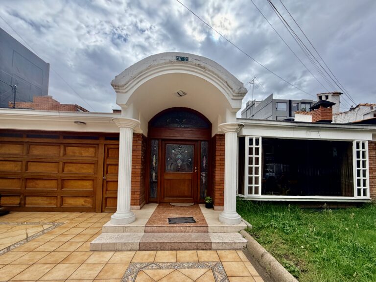 Fachada frontal de la casa Funsinapsis de un piso con un porche de entrada blanco en forma de arco apoyado sobre dos columnas. Cuenta con una puerta principal de madera con vitrales, un portón de garaje de madera a la izquierda y un gran ventanal con un pequeño jardín a la derecha.